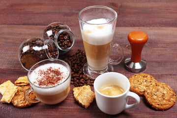 Cups of espresso, latte and cappuccino, crackers, holder with ground coffee, tamper and coffee beans on wooden table.