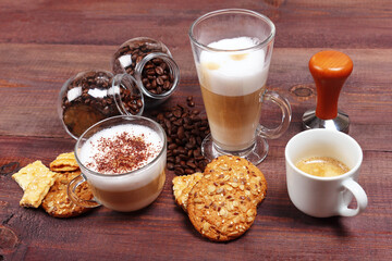 Cups of espresso, latte and cappuccino, crackers, cookies, holder with ground coffee, tamper and cans of coffee beans on wooden table.