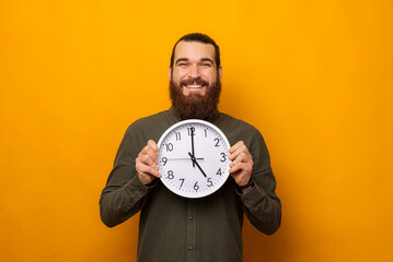 Handsome bearded man is holding a round white wall clock while smiling.