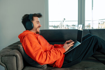 Handsome man laughing while using laptop with headset sitting on sofa in living room. Remote...