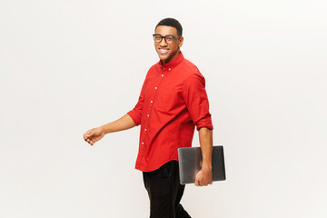 Cheerful handsome smiling African-American young man in red shirt carrying laptop and walking...