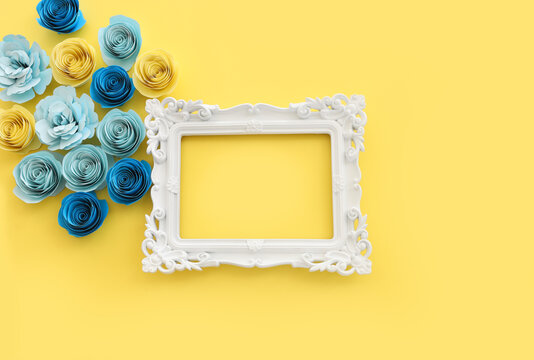 Top view image of paper flowers composition and empty white photo frame over yellow pastel background
