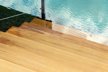 Close-up detail view of natural wooden stair steps of larch ladder into clean blue water of lake, sea or pond. Waterpoof timber material. Damp-proof striped decking board terrace surface - Powered by Adobe