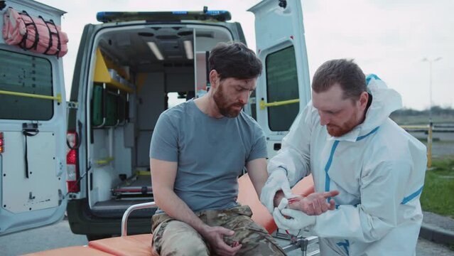 Shot of field doctor rewinding hand bandage of military man in uniform wounded by artillery fire during combat. Care of health of wounded soldier. Medic treating wounds of injured