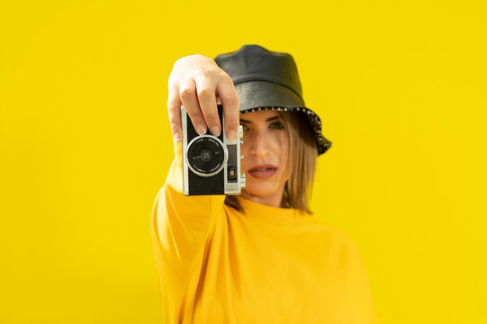 A young caucasian woman in a black hat holding a vintage photo camera over a yellow colored wall