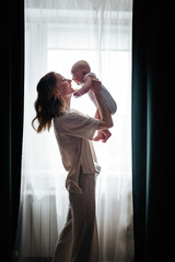 Young woman holding baby girl in her hands and looking at her with love while standing in front of the window.