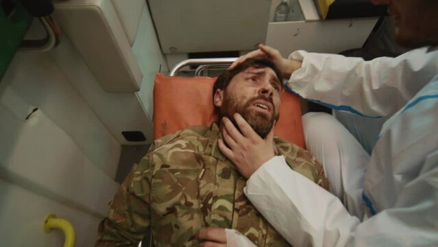 Close-up shot of bearded man lying on medical trolley for transportation, suffering in pain. Doctor taking care of injured. Medic helping badly wounded soldiers to survive. High quality of treatment.