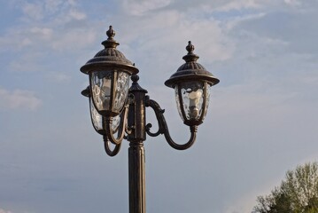 Fototapeta na wymiar black iron pole with three lanterns and lamps on the street against the blue sky and clouds