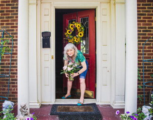 Pretty Senior Woman in shorts and tee-shirt at door to brick house with pillars and sunflower...