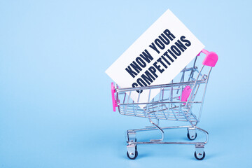 A white card with text KNOW YOUR COMPETITIONS stands in a shopping trolley on blue background.