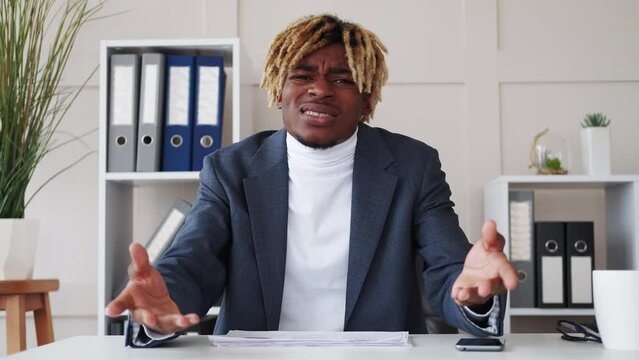 Work quarrel. Arguing black man. Online meeting. Angry outraged guy sitting desk expressive explaining his point of view in light room interior.