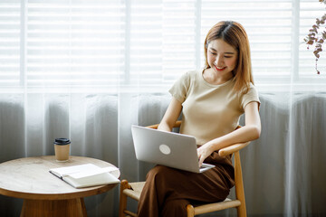 Young Asian lady studying online from home, reading a book and using laptop pc, sitting on the coffee shop, free space. E-learning, web-based education concept