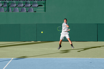 Professional tennis player performs forehand hit on hard tennis court. Young male athlete with tennis racket in action. Junior tennis sport. Green background, banner, copy space - obrazy, fototapety, plakaty