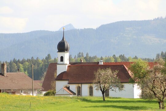 Kirche in einem kleinen Dorf im Allgäu, Berge im Hintergrund. Bäume blühen im Frühling.