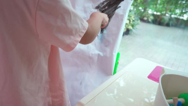 Close up of painting process so dirty at home.Asian child girl standing and paint on white canvas on an easel in playroom.Try to use paint brush with black color. Children's creativity. 