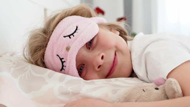 girl sleeping. A beautiful, sweet little girl with long hair is lying in bed hugging a soft toy rabbit. The eyes are covered with a sleep mask. The child woke up, took off his mask and smiled a happy 