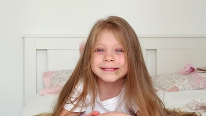 A beautiful, sweet little girl with blue eyes and long hair lies on her stomach in bed and smiles. The child is naughty and indulges. Children's pranks and games in the bed.