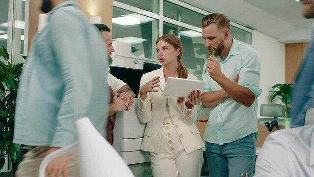 Two bearded men in their work suits are talking to a blonde woman holding a tablet in her white suit, they re in the office conversing about their work