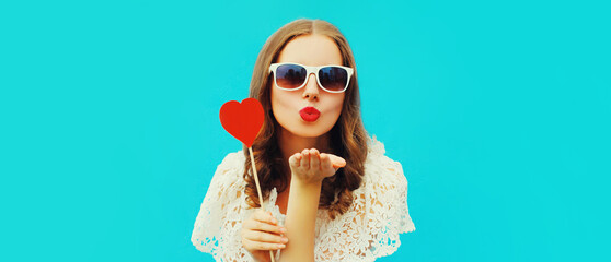 Portrait beautiful young woman with red heart shaped lollipop blowing lips sending sweet air kiss on blue background