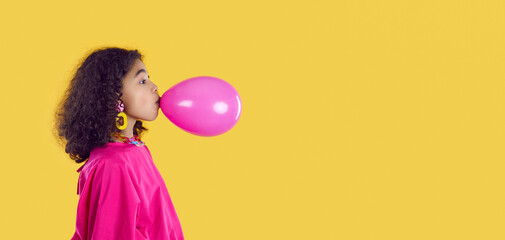 Funny stylish dark skinned preteen girl inflates pink balloon on yellow background. Profile portrait of ethnic curly little girl who inflates balloon before children's party. Copy space. Web banner. © Studio Romantic