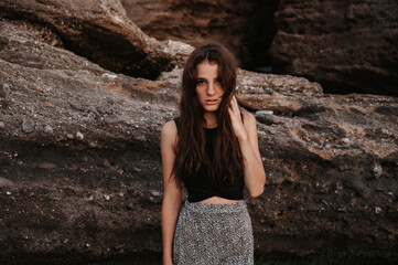 A beach photo of a beautiful sensual young woman with long dark hair in a dress posing on the beach by large boulders.