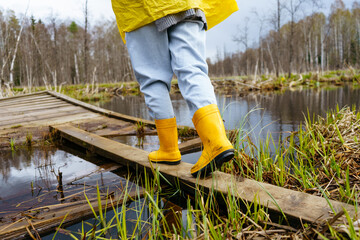 The legs of a woman in yellow boots walk on a wooden bridge near a lake in the forest