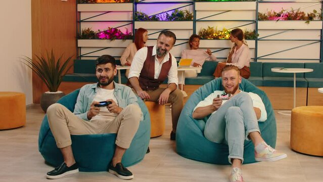 Two young handsome men with beards are sitting in bean bag chairs and playing video games while an older man behind them is watching them play and in the fat back three women are sitting on the sofa