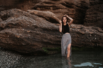 A picture of a beautiful sensual young woman with long dark hair in a dress posing on the beach by large boulders.