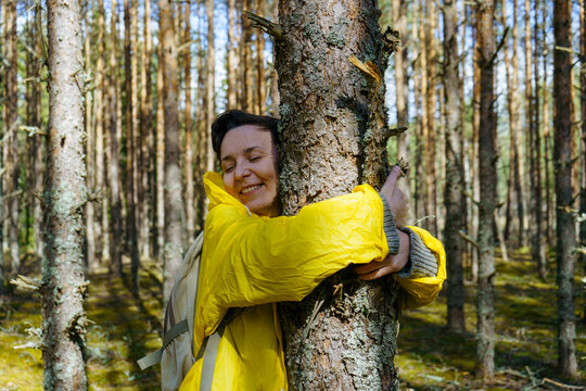 A young woman in a yellow raincoat hugs a pine tree, closes her eyes and smiles