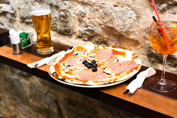 Delicious pizza with prosciutto and mushrooms, a glass of beer and a glass of aperol at outdoor...