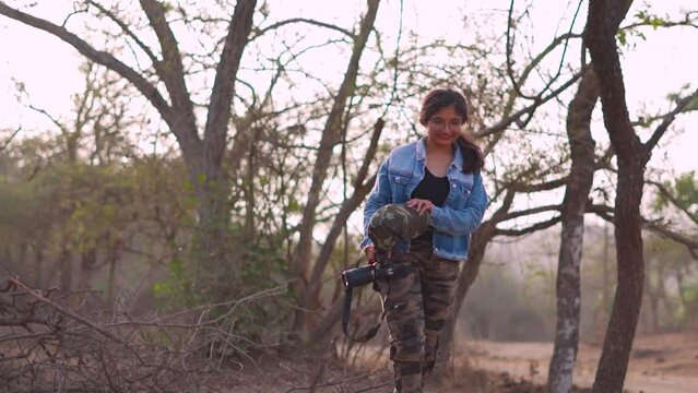 Slow motion portrait of beautiful Indian female holding camera and cap in hand walking at Forest of Sasan Gir, Gujarat, India. Confident female photographer. 