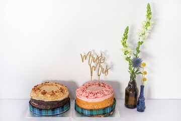 Two delicious cakes, one chocolate with caramel or buttercream frosting, the other a classic sponge base with strawberry frosting and a wedding cake golden topper decoration that reads "Mrs & Mrs"