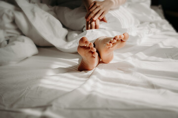 Woman is lying with bare feet in bed, sleeping under white cosy and comfortable bedclothes. Female feet on bed. Good morning, woman sexuality concept, self-care and relaxation, resting concept.