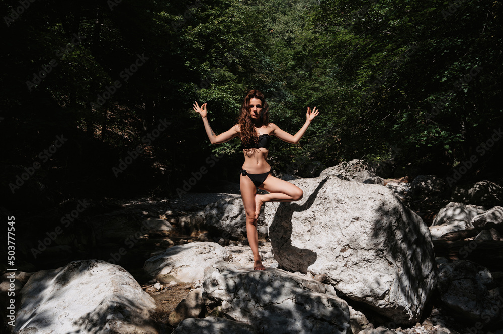 Wall mural young sexy brunette girl in a black swimsuit stands in a yoga pose on the rocks by the river in natu