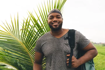Foto op Plexiglas Half length portrait of cheerful male traveller in casual bandanna posing near tropical palm trees enjoying summer vacations for recreation, happy African American hipster guy with touristic backpack © BullRun