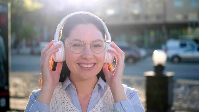 An attractive young brunette woman with glasses and white headphones listens to music on the street during the day and smiles