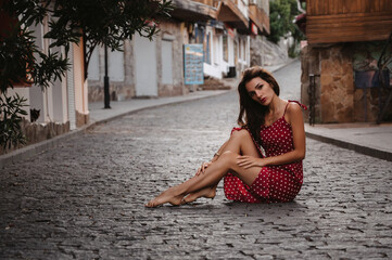 Sexy young Caucasian girl in a red dress posing sitting on a paving stone on a beautiful European...