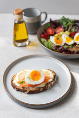 Healthy breakfast or snack with medium boiled eggs on toasted bread with cheese cream, salmon and lemon slices on grey plate. Bowl of salad and olive oil nearby