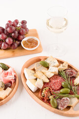 Vertical composition with antipasti plate - variety of cheeses, sausages served with glass of wine, sun dried tomatoes, olives, jams and herbs on white table. Cheese and meat snacks platter