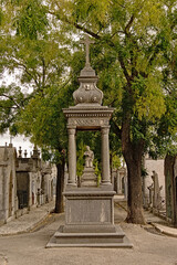 Fototapeta premium Shrine with statue of a kneeling women in a lane with grave chapels and trees in Alto de Sao Joao cemetery, Lisbon, Portugal 