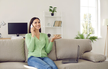 Portrait of smiling happy young woman taking break from work talking to friend on mobile phone. Woman working remotely at home is sitting in living room on sofa next to laptop and talking on phone.