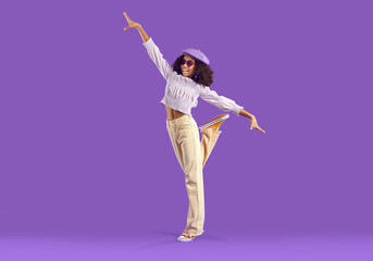 Happy kid poses in trendy casual outfit. Beautiful child having fun in fashion studio. Pretty Black girl wearing purple beret, lilac top and beige denim pants standing on one foot on purple background