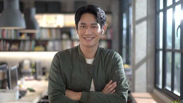 Selective focus front view of a young handsome Asian businessman in a jacket wearing wireless earbuds standing with arms crossed while looking and smiling at the camera in a blurred modern office.