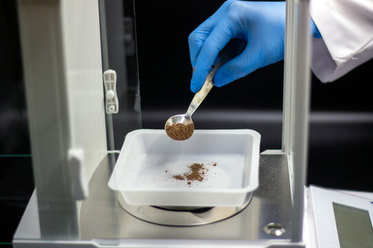 The scientist is weighing the sample. Digital analytical balance. Close up scientist's hands in blue gloves. Brown powder sample in the metal spatula. Laboratory concept. 