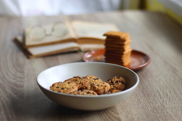 Bowl of chocolate chip cookies, plate of biscuits, open books and reading glasses on the table. Selective focus.