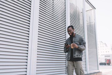 Fototapeta premium Millennial black tourist smiling in city using cellular application for tracking location GPS during travel walking, cheerful hipster guy in optical eyewear enjoying online networking and chatting