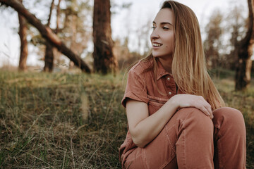 Obraz premium Happy smily young blonde woman in jumpsuit siiting on glade in park looking sideways and smiling 