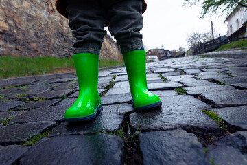 A small child in green rubber boots stands on a paved road.