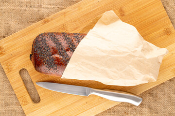 One sweet homemade poppy seed roll in a paper bag, with a knife on a bamboo cutting board, close-up, top view.