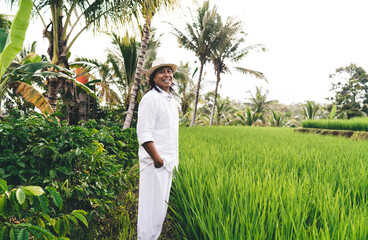Cheerful Balinese entrepreneur in hat and white clothes laughing while visiting own farmland with rice fields and coffee bushes - enjoying business lifestyle at countryside of Indonesia, happiness